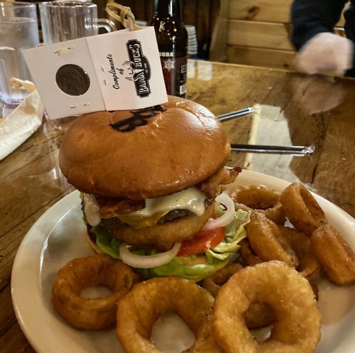 A hearty burger with bacon, cheese, lettuce, tomato, and onion, served with a side of onion rings on a plate.