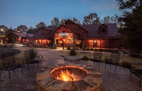 A cozy log cabin lodge at dusk, featuring a fire pit and outdoor seating in a serene landscape.