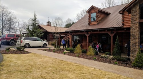 A rustic log cabin building with a gravel path, surrounded by trees and people enjoying the outdoor space.