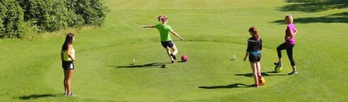 Four young women are playing soccer on a grassy field, with one kicking a ball while the others watch.