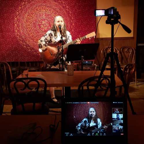 A singer performs with a guitar on stage, captured from a camera view on a laptop, with a decorative backdrop.
