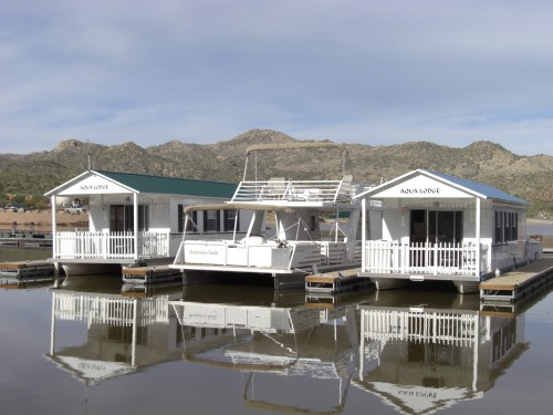 Three white houseboats with green roofs docked on calm water, reflecting mountains in the background under a cloudy sky.