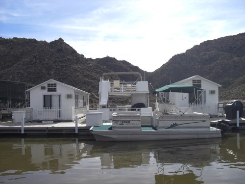 A houseboat docked near white houses on a calm lake, surrounded by rocky hills under a clear sky.