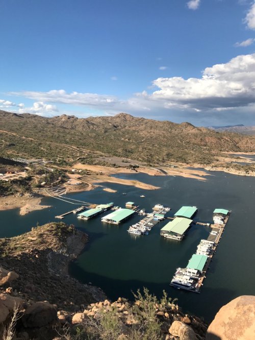 Aerial view of a marina with green-roofed boats on a calm lake, surrounded by hills and a blue sky with clouds.
