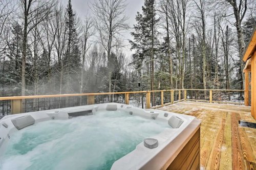A hot tub on a wooden deck surrounded by snowy trees, with steam rising in a serene winter landscape.
