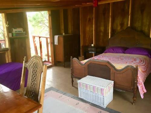 Cozy wooden cabin interior with a bed, a small table, and a wicker storage box, featuring purple bedding and rustic decor.