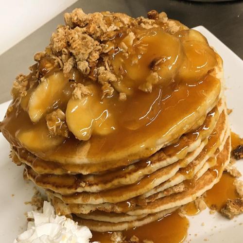 A stack of pancakes topped with caramel sauce, sliced bananas, crushed nuts, and a dollop of whipped cream.