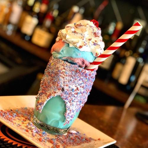 A colorful milkshake topped with whipped cream, a cherry, and sprinkles, served in a glass with a striped straw.
