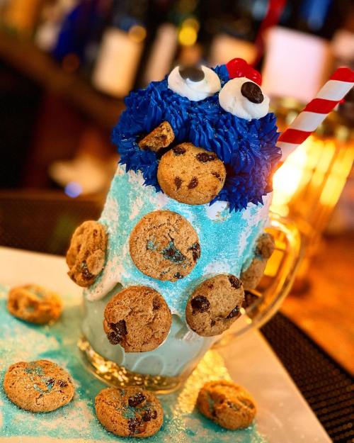 A colorful milkshake topped with blue frosting, a cookie monster face, and surrounded by chocolate chip cookies.