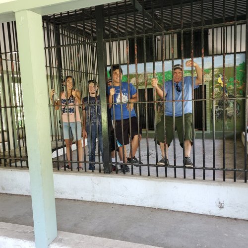 Four people stand behind bars in a cage-like structure, smiling and posing for the camera. Colorful murals are visible behind them.