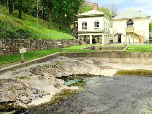 A serene landscape featuring a stone building by a calm water body, surrounded by greenery and a rocky shore.