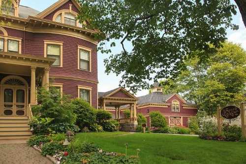 A charming Victorian-style house with purple siding, surrounded by lush greenery and colorful flower beds.