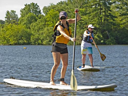 Two women paddleboarding on a calm lake, surrounded by greenery, wearing life jackets and enjoying the sunny day.