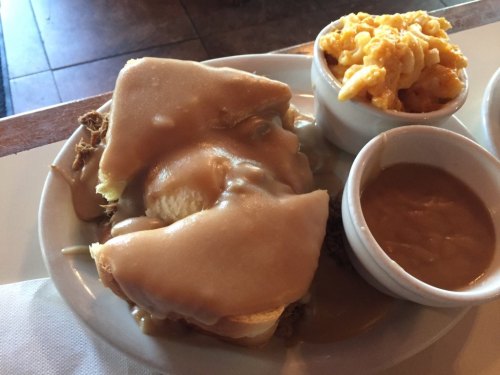 A plate with a sandwich topped with gravy, alongside macaroni and cheese and a small bowl of beans.