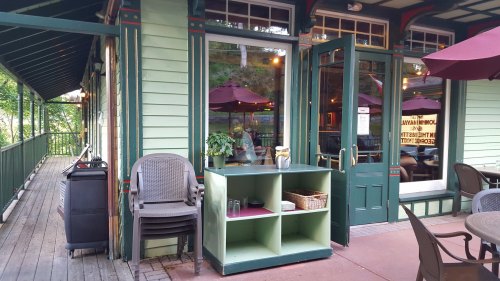 A charming green building with a porch, featuring tables, chairs, and umbrellas, inviting guests to relax outside.
