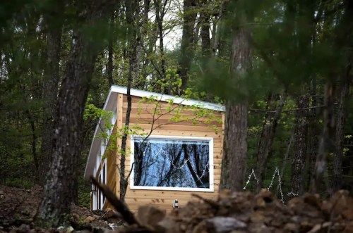 A small wooden cabin nestled among trees, reflecting the surrounding forest in its large window.