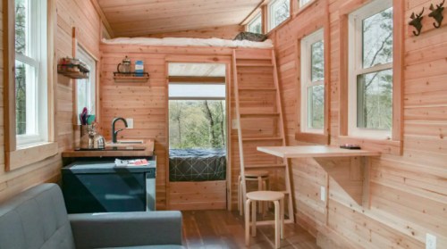 Cozy tiny house interior with wooden walls, a kitchenette, a loft bed, and a small dining area by the windows.