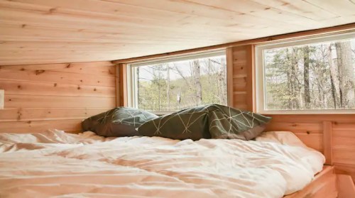 Cozy bedroom with wooden walls, a large bed, and two windows overlooking a forested area.