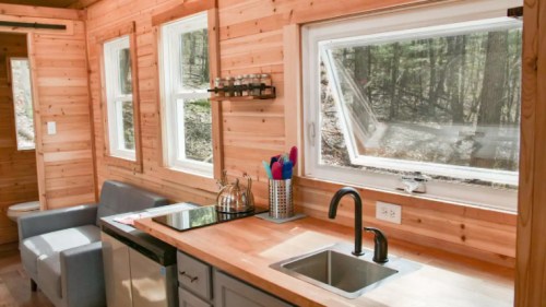 Cozy wooden kitchen with a sink, stove, and large windows overlooking a forested area.