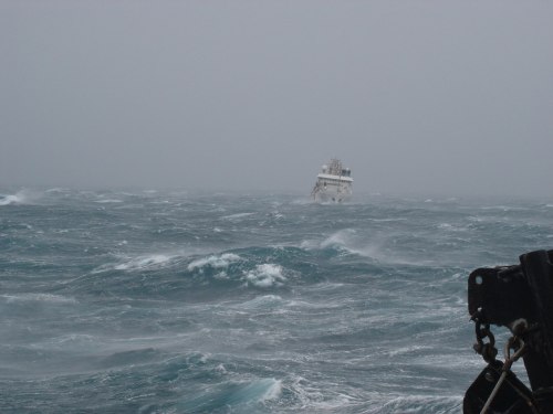 A ship struggles against rough seas and strong winds in a stormy, overcast atmosphere.