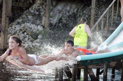 Two children slide into the water from a dock, splashing as they enjoy a summer day. A lifeguard watches nearby.