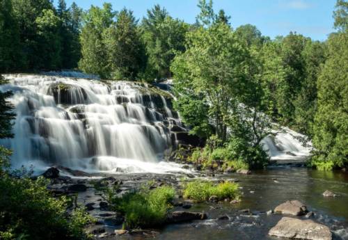 A serene waterfall cascades over rocks, surrounded by lush green trees and a clear blue sky.