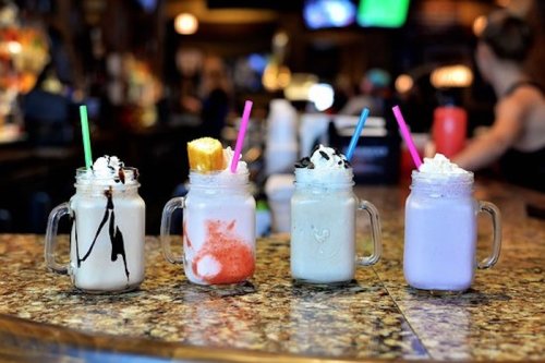 Four colorful milkshakes in mason jars with whipped cream and straws, displayed on a bar counter.