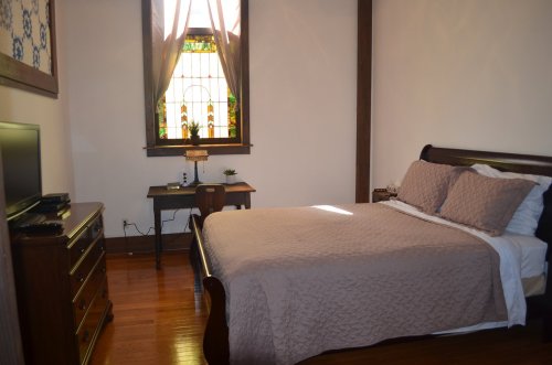 Cozy bedroom with a wooden bed, dresser, desk, and a stained glass window, featuring warm natural light.