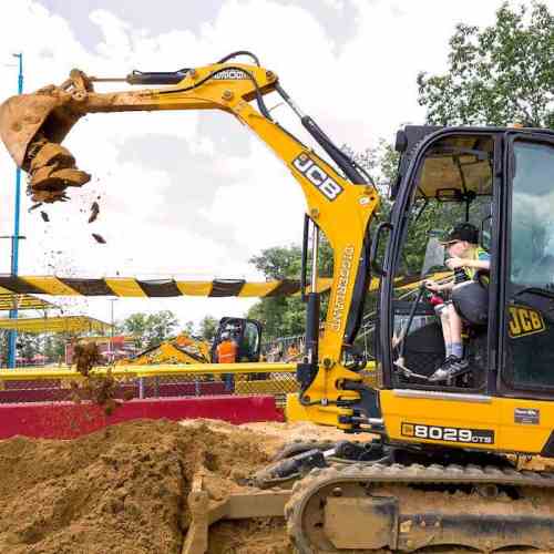 A child operates a yellow JCB excavator, lifting a scoop of dirt at a construction-themed play area.