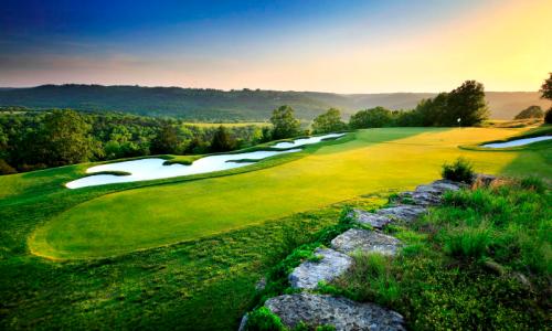 A scenic golf course at sunset, featuring lush green fairways and sand traps, surrounded by rolling hills and trees.