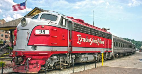 A vintage red and silver train with "Bromley Central Railway" on the side, parked near a station with an American flag.