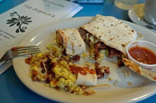 A plate with a partially eaten breakfast burrito, scrambled eggs, bacon, and a side of salsa on a blue table.
