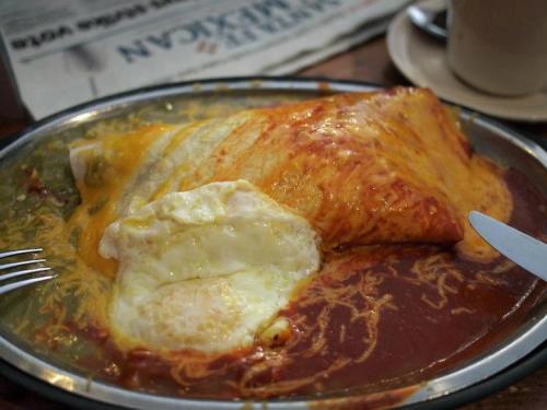A plate of cheesy enchiladas topped with red sauce and a fried egg, served with a fork and knife.