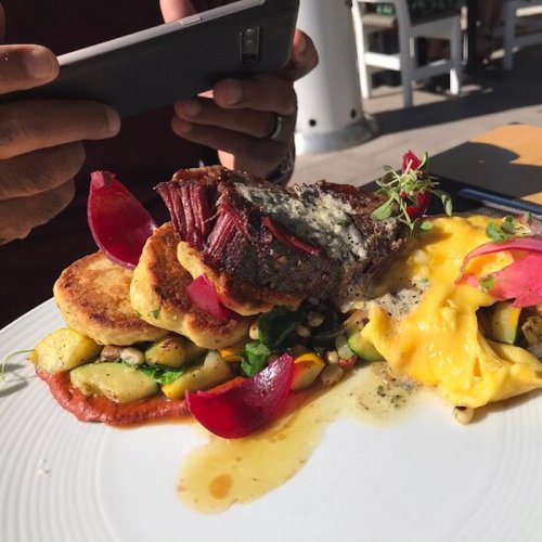 A plate of gourmet food featuring steak, vegetables, and garnishes, with a person taking a photo in the background.