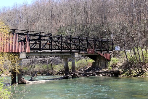 An old, partially collapsed bridge spans a clear river, surrounded by bare trees and rocky banks in a serene landscape.