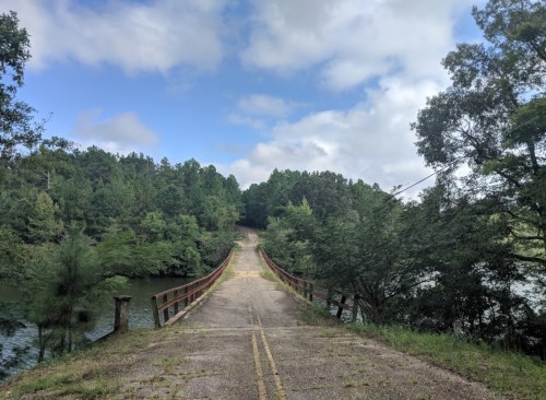 A narrow, weathered road leads to a bridge over a calm body of water, surrounded by lush green trees and a cloudy sky.