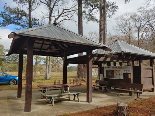 A wooden pavilion with a metal roof, picnic tables, and a trail map, surrounded by trees in a park setting.