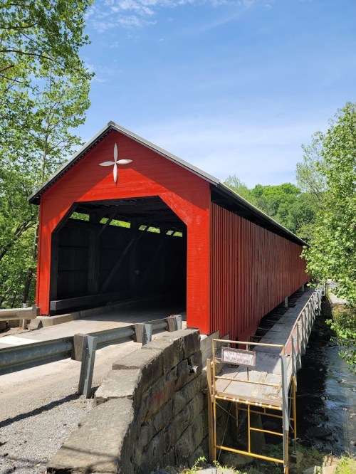A red covered bridge spans a small stream, surrounded by green trees and a clear blue sky.