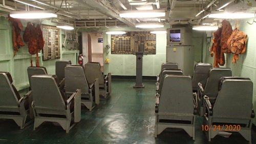 Interior of a ship's control room with rows of seats, safety gear on the walls, and a monitor displaying information.