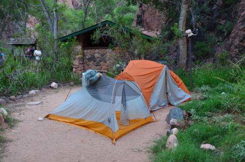 Two tents set up in a grassy area surrounded by trees and rocks, with a stone building in the background.