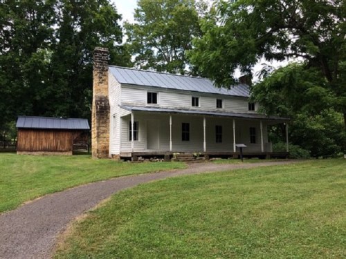 A white two-story house with a metal roof, surrounded by greenery and a gravel path, with a wooden shed nearby.