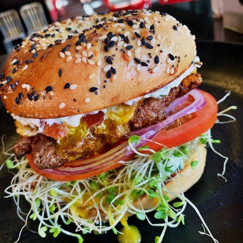 A close-up of a sesame seed bun burger with fried egg, crispy chicken, tomato, onion, and sprouts on a black plate.