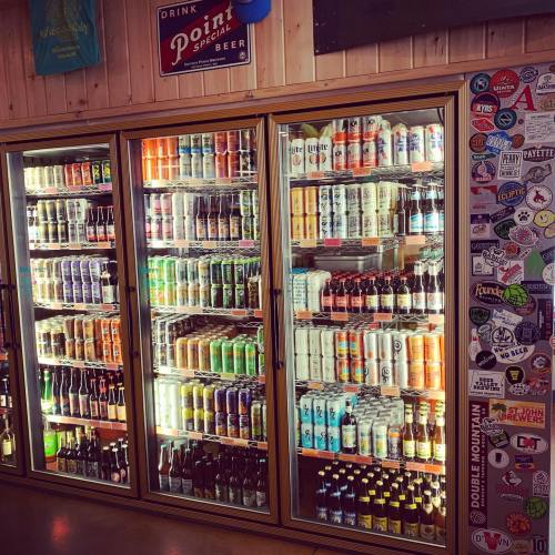 A display of various canned and bottled beverages in a glass-fronted cooler, with colorful labels and a sticker-covered wall.