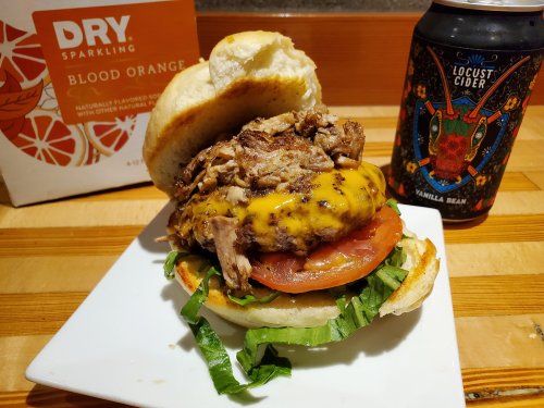 A close-up of a burger with grilled chicken, cheese, lettuce, and tomato on a plate, alongside a can of cider.