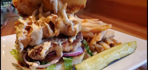 A towering burger with lettuce, onion, and sauce, served with fries and a pickle on a white plate.