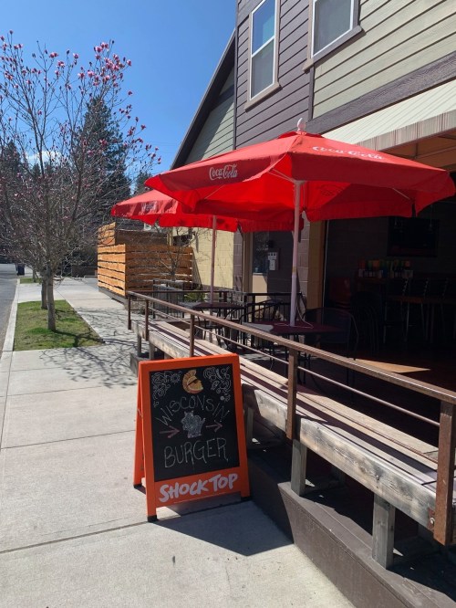 A sunny outdoor dining area with red umbrellas and a chalkboard sign advertising "Wisconsin Burger" and "Shock Top."