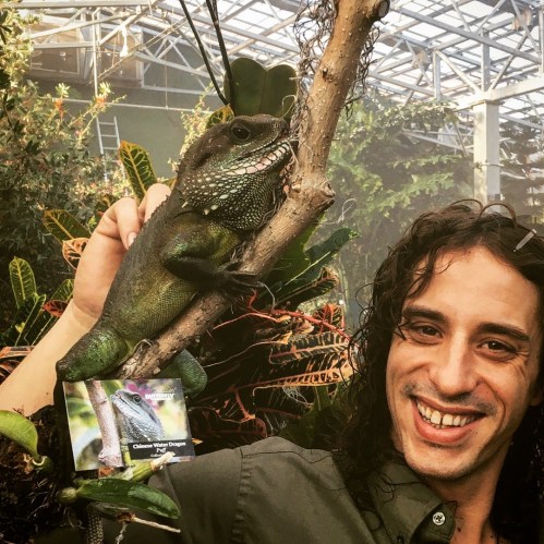 A person with curly hair poses next to a green lizard on a branch in a lush greenhouse setting.