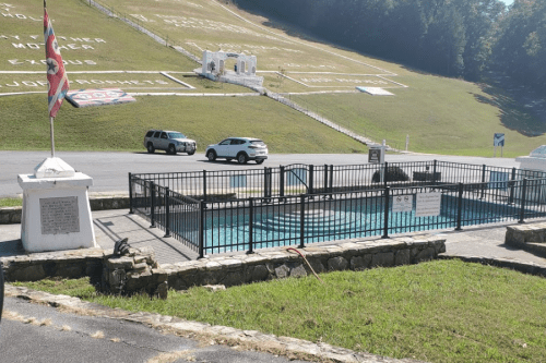 A fenced swimming pool area with a flag nearby, set against a hillside with white inscriptions.