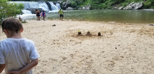 A sandy riverbank with a waterfall in the background; children play near the water while an adult stands nearby.