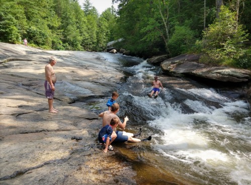 A group of people enjoying a rocky riverbank, with two children playing in the water and an adult sliding down a rock.
